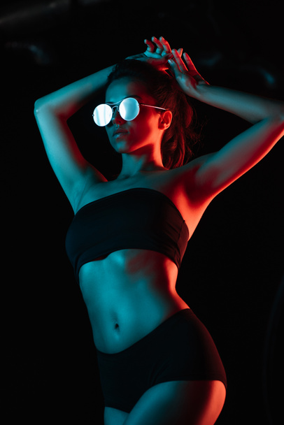 trendige junge Frau mit Sonnenbrille, die die Haare isoliert auf schwarz berührt  - Foto, Bild