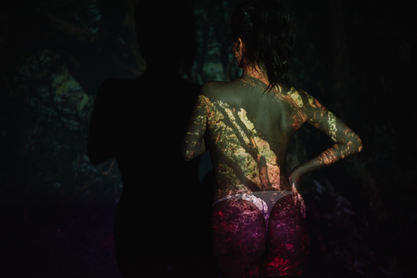 Вид сзади девушки с тенями на теле, стоящей с рукой на бедре на черном
  - Фото, изображение