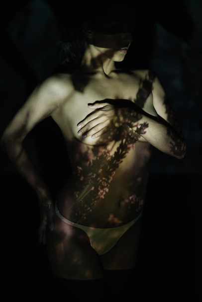 περικομμένη θέα της νεαρής γυναίκας που καλύπτει τα στήθη, ενώ στέκεται με το χέρι στο ισχίο σε μαύρο με σκιές  - Φωτογραφία, εικόνα