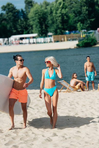 przystojny młody mężczyzna trzyma Surfboard podczas spaceru w pobliżu atrakcyjnej kobiety w strój kąpielowy  - Zdjęcie, obraz