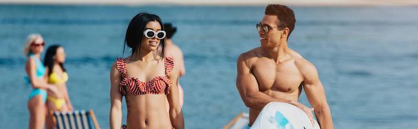 panoramic shot of handsome man holding surfboard near attractive woman in swimsuit - Photo, Image