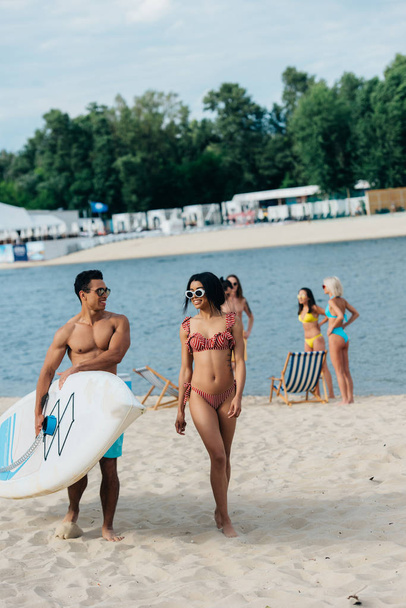 alegre mixto raza hombre celebración surf tablero cerca africano americano chica en traje de baño
 - Foto, Imagen