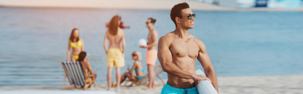 plano panorámico de hombre de raza mixta guapo sosteniendo tabla de surf mientras amigos multiculturales descansando en la playa
 - Foto, Imagen