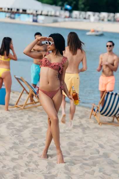 beautiful african american woman in swimsuit looking away while standing on beach near multicultural friends - Фото, изображение