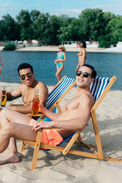 glimlachende multiculturele vrienden die naar de camera kijken terwijl ze op het strand zitten in chaise lounges en flessen met bier houden - Foto, afbeelding