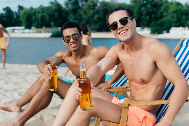 handsome multicultural friends smiling at camera while sitting in chaise lounges and holding bottles with  beer - Foto, Imagem