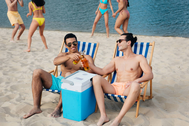 cheerful multicultural friends clinking bottles of beer while sitting in chaise lounges near portable fridge on beach - Photo, Image