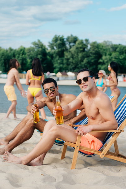 przystojny wielokulturowych przyjaciół trzymając butelki piwa i uśmiechając się do kamery siedząc w szezlongi na plaży - Zdjęcie, obraz