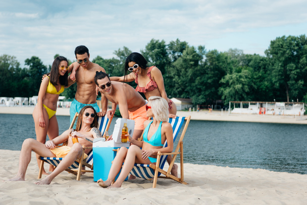 χαρούμενοι πολυπολιτισμικοί φίλοι στέκονται κοντά σε άντρα και γυναίκα που κάθονται σε σεζλόνγκ με μπουκάλια μπύρας - Φωτογραφία, εικόνα