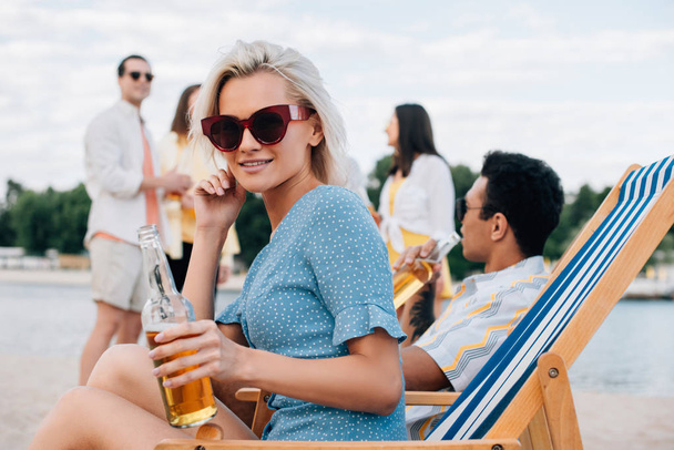 gyönyörű nő napszemüveg gazdaság üveg sört ülve a hintó Lounge közelében multikulturális barátok - Fotó, kép