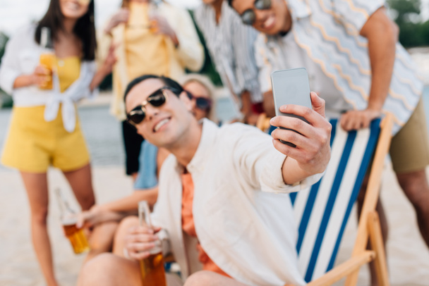 foco seletivo de jovem alegre tomando selfie com amigos multiculturais se divertindo na praia
 - Foto, Imagem