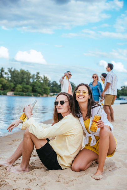 Wesoła Młoda para siedzi na piasku i trzyma butelki piwa w pobliżu wielokulturowych przyjaciół - Zdjęcie, obraz