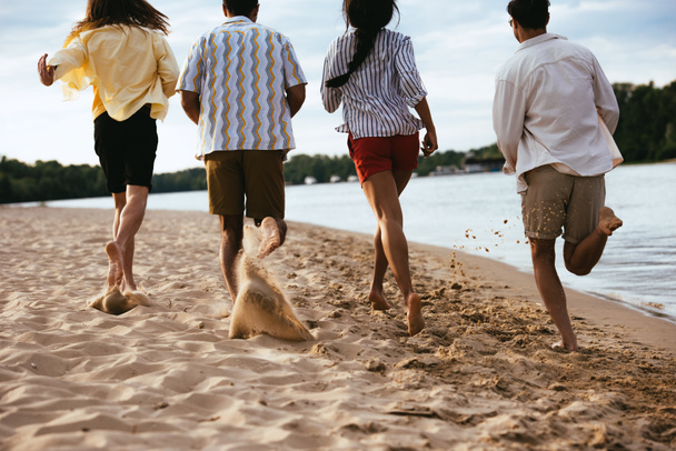 vista trasera de amigos multiétnicos corriendo en la playa de arena cerca del río
 - Foto, Imagen