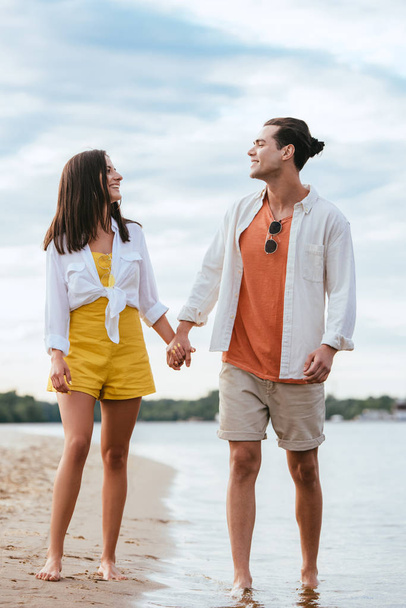 cheerful young couple holding hands and looking at each other while walking on beach - Foto, imagen