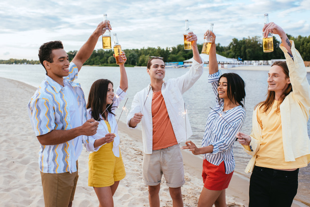cheerful multicultural friends holding bottles of beer in raised hands and lighting sparklers on beach - Photo, Image