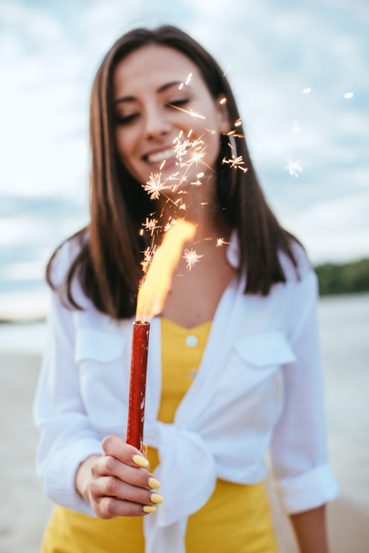 selective focus of happy young woman holding sparkler while standing on beach - Photo, Image