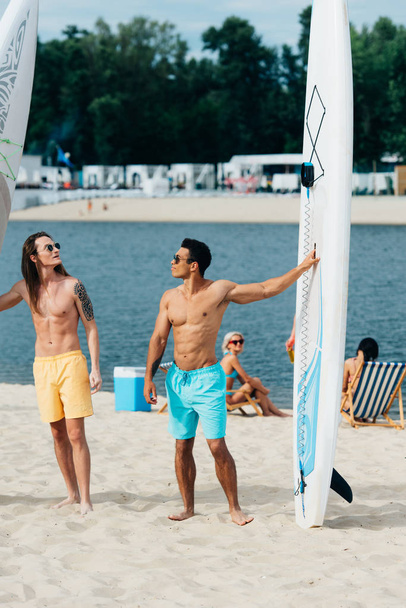 bello, giovani uomini multiculturali in piedi sulla spiaggia vicino tavole da surf
 - Foto, immagini
