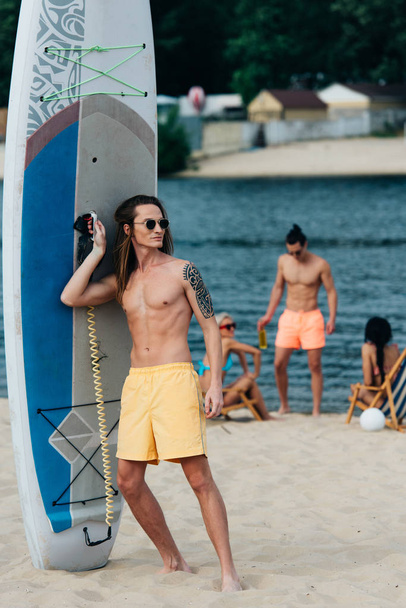 pohledný mladý muž, který se dívá, zatímco stojí nedaleko surfového prkna na pláži - Fotografie, Obrázek