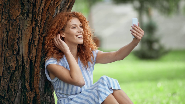χαρούμενο κοκκινομάλλα κορίτσι αγγίζοντας το πρόσωπο ενώ παίρνετε selfie  - Πλάνα, βίντεο
