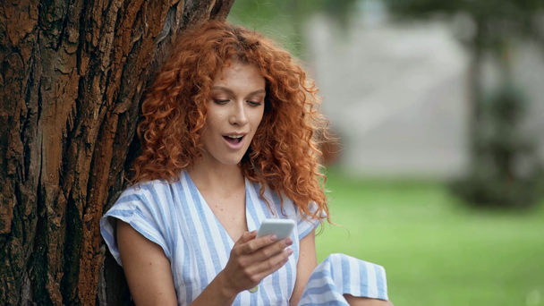 surprised redhead girl using smartphone in park  - Footage, Video