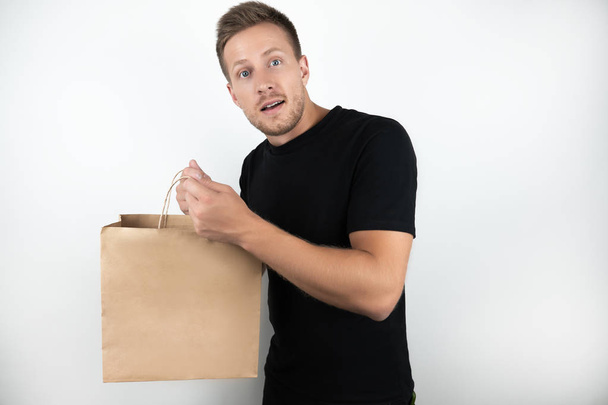 handsome young man wearing black t-shirt holding shopping craft papper bag with goods returning from supermarket on isolated white background - Fotografie, Obrázek