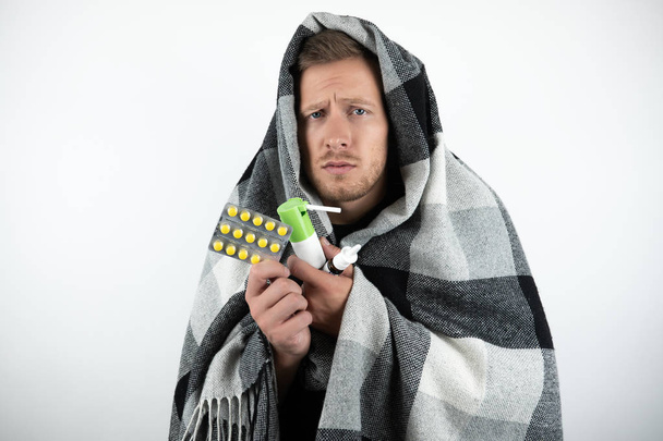 handsome young sick man wrapped checkered plaid holds pills and nasal spray looks pityingly on isolated white background - Foto, Bild