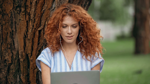 curly woman using laptop and listening music in park  - Video