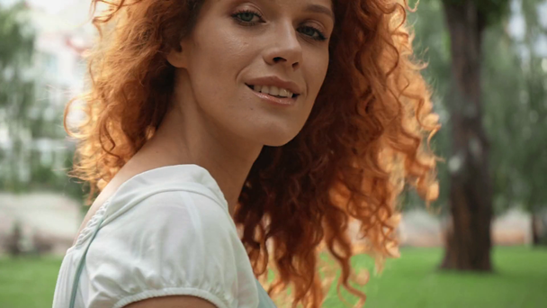 χαρούμενο κοκκινομάλλα κορίτσι αγγίζοντας τα μαλλιά και το περπάτημα στο πάρκο  - Πλάνα, βίντεο
