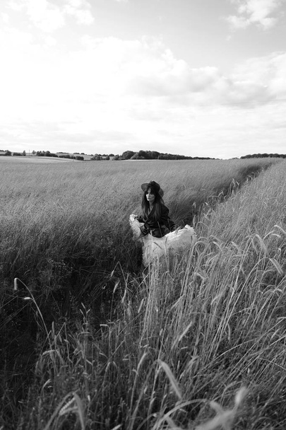 Retrato preto e branco de bela jovem mulher em jaqueta de couro, chapéu preto, vestido branco., andando na grama
.  - Foto, Imagem