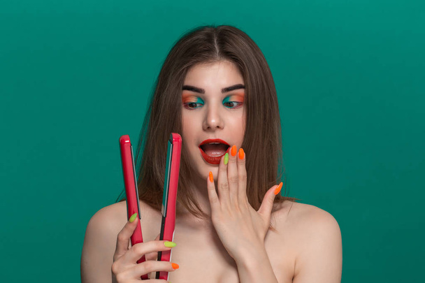 Portrait of beautiful young woman with bright color make-up doing hair style herself with a purple straightening iron. - Photo, Image
