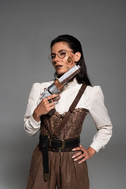 attractive steampunk woman standing with hand on hip and holding pistol isolated on grey - Photo, Image