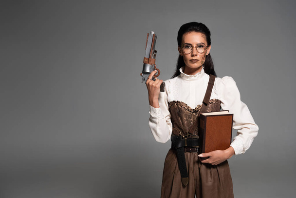 front view of steampunk woman holding book and vintage pistol on grey - Foto, Imagen