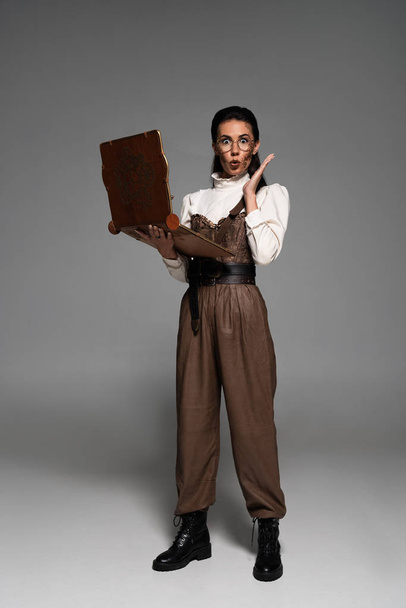 full length view of shocked steampunk woman in glasses using vintage laptop on grey - 写真・画像