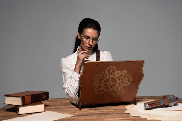 Žena v brýlích s dokumenty a se starožitným notebookem na pracovišti izolovaný na šedé - Fotografie, Obrázek