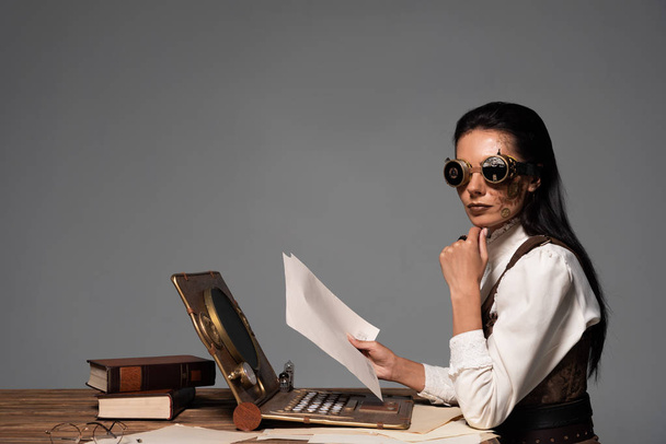 mulher steampunk segurando documentos à mesa com laptop vintage isolado em cinza
 - Foto, Imagem