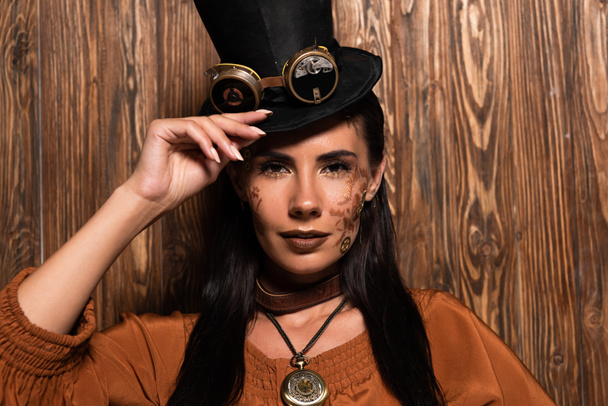 front view of steampunk woman touching top hat with goggles looking at camera on wooden - Foto, afbeelding