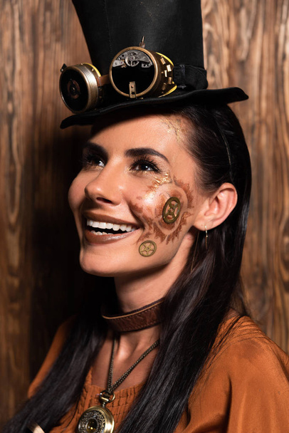 cheerful steampunk woman with makeup looking up and smiling on wooden - Photo, Image