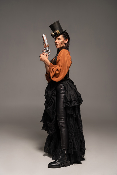 full length view of steampunk woman in top hat with goggles holding pistol on grey - Photo, Image