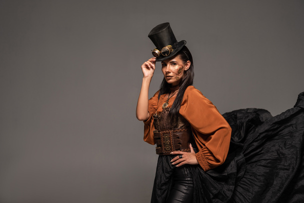 steampunk woman in top hat with goggles standing with hand on hip isolated on grey - Photo, Image