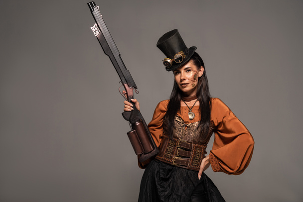 attractive steampunk woman in top hat holding gun and standing with hand on hip isolated on grey - Photo, Image