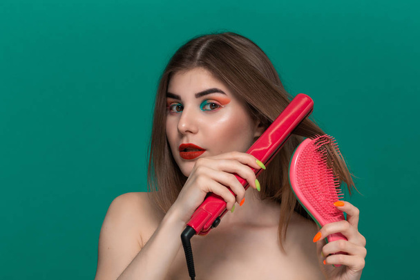 Portrait of beautiful young woman with bright color make-up doing hair style herself with a purple straightening iron and a hairbrush. - Foto, imagen