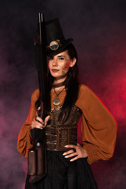 attractive steampunk woman holding gun and standing with hand on hip in pink smoke - Photo, Image