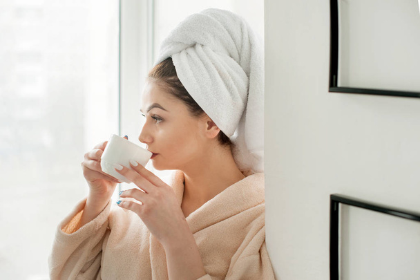 Rapariga com uma toalha na cabeça está bebendo com cofee copo, perto peitoril da janela no banheiro
 - Foto, Imagem
