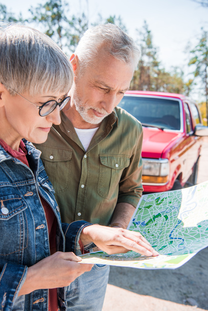 senior couple of tourists standing near car and looking at map in sunny day - Photo, image