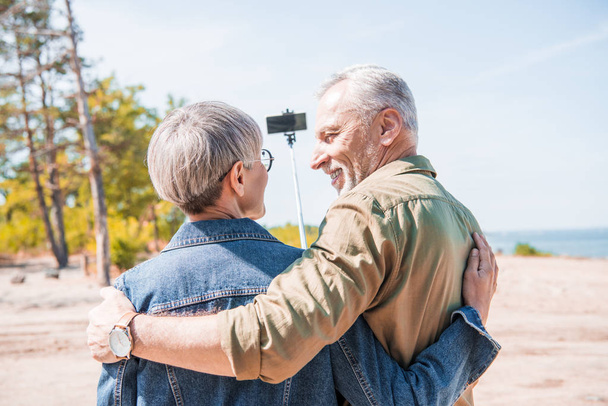 πίσω όψη του ζευγαριού ηλικιωμένων Αγκαλιάζοντας και παίρνοντας selfie στην ηλιόλουστη μέρα - Φωτογραφία, εικόνα