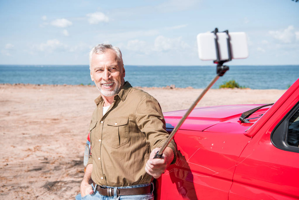 sonriente hombre mayor de pie cerca de coche rojo y tomar selfie en día soleado
 - Foto, imagen