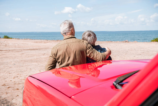 задний вид пожилой пары, стоящей рядом с красной машиной на пляже и обнимающейся в солнечный день
 - Фото, изображение