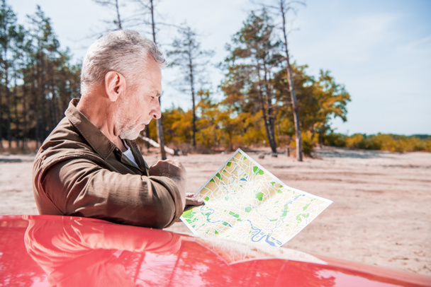 turista senior centrado de pie cerca de coche rojo y mirando el mapa en el día soleado
 - Foto, Imagen