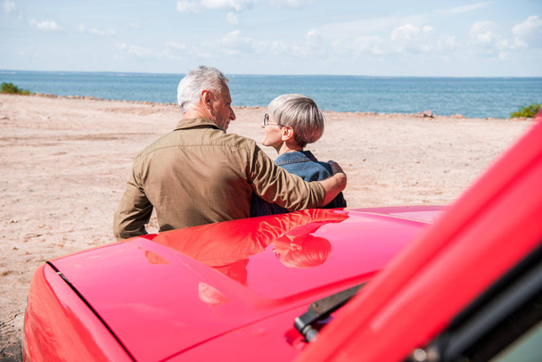 ビーチで赤い車の近くに立って、晴れた日に抱き合うシニアカップルのバックビュー - 写真・画像