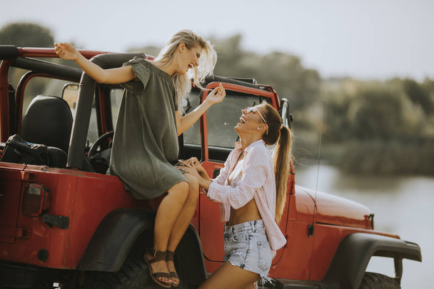 zwei junge Frauen amüsieren sich an einem heißen Sommertag auf der Motorhaube eines Autos am See - Foto, Bild